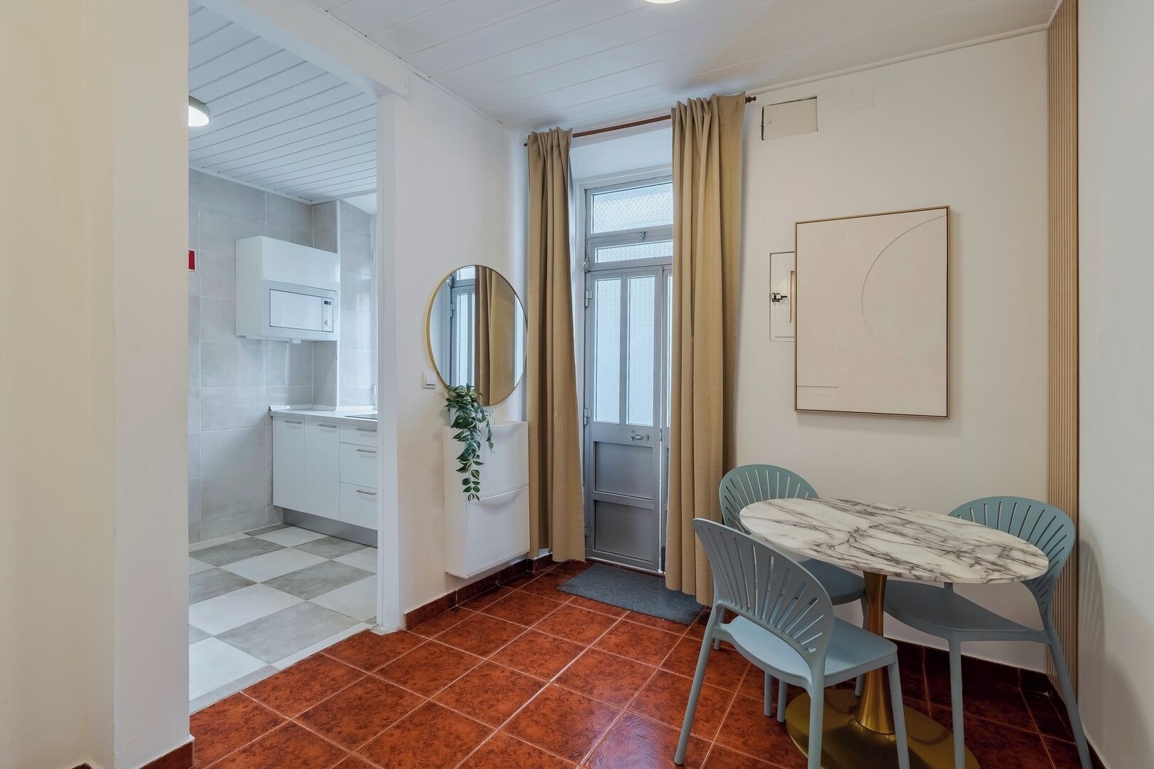 A compact room with a checkered floor, a small dining table with chairs, a mirror, and a window with curtains.
