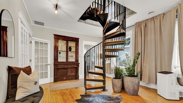 Un salón amplio con una escalera espiral, suelo de madera, una gran planta en maceta y un moderno equipo de aire acondicionado.