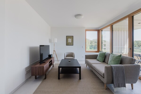 Una sala moderna con televisor de pantalla plana, una mesa de centro, un sofá con cojines, una lámpara de pie y ventanas amplias que ofrecen vistas al exterior.