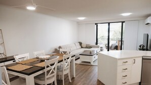 Una cocina moderna con armarios blancos, una zona de comedor con mesa y sillas, y una sala de estar con sofá y televisor.