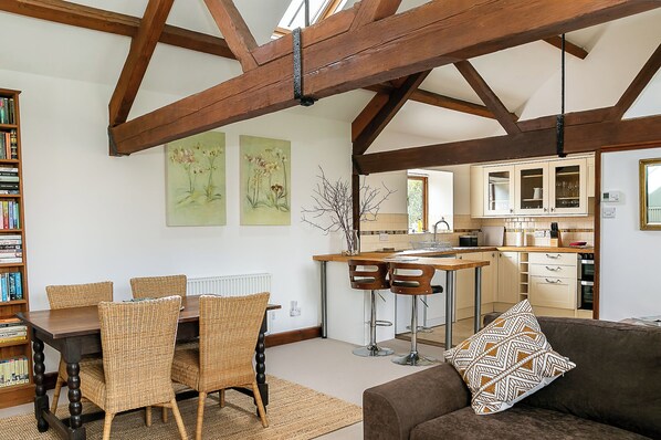 A spacious living area with a dining table, wicker chairs, a sofa with a patterned cushion, and a kitchen in the background.