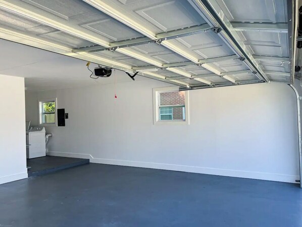 Un garage avec un sol en béton, un plafond équipé de néons, un lave-linge et une petite fenêtre donnant sur l’extérieur.