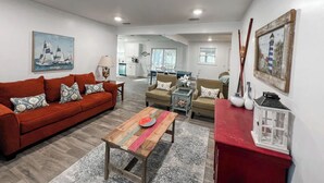 A living room with a red sofa, a wooden coffee table, and a red cabinet. There is a painting of sailboats on the wall and a lamp on a side table. - Cozy Coastal Abode- 4bed,3bath (Brunswick)