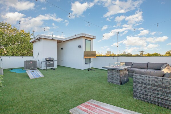 Eine Dachterrasse mit Kunstgrashalm, einem kleinen Gebäude, Gartenmöbeln und Lichterketten.