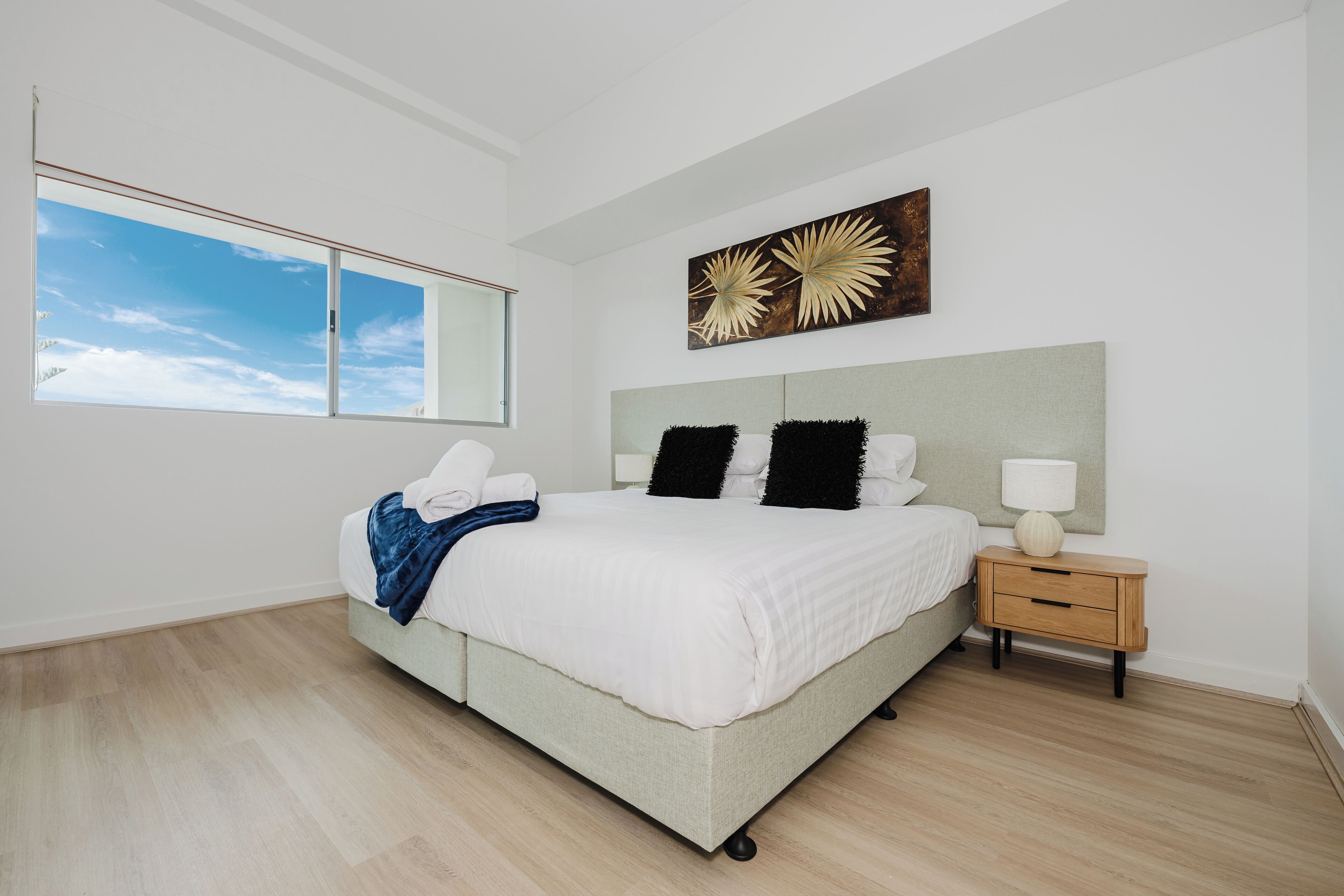 A modern bedroom with a large bed, a nightstand, a wall-mounted artwork, and a window with a view of the sky.