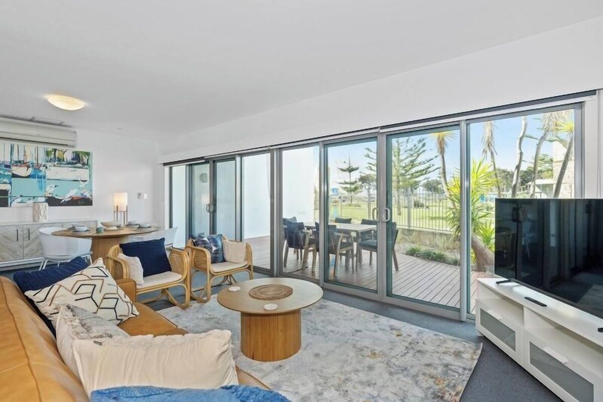 A modern living room with a large TV, comfortable seating, and a glass door leading to an outdoor patio.