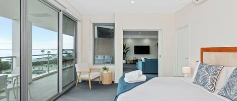 Moderni hotellihuone, jossa on suuri sänky, parvekkeelta avautuva näköala, oleskelualue ja televisio huoneen nurkassa.