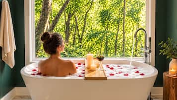 A woman in a bathtub with rose petals and candles, a window with a forest view, and a wooden stool with a candle.