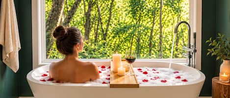 A woman in a bathtub with rose petals and candles, a window with a forest view, and a wooden stool with a candle.