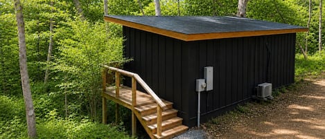 Eine kleine, dunkelbraune Holzhütte mit einer Holztreppe, umgeben von dichtem Grün.