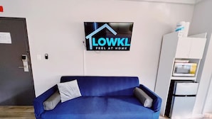 Ein modernes Wohnzimmer mit einem blauen Sofa, einem an der Wand befestigten Schild mit der Aufschrift "LOWKL FEEL AT HOME" und einer kleinen Küchenzeile mit Kühlschrank und Mikrowelle.