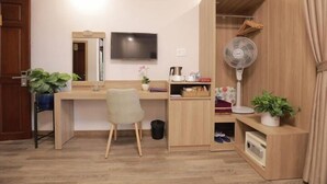 Een kleine kamer met een houten bureau, een stoel, een televisie, een ventilator en een klein plankje met persoonlijke spullen.