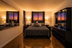 A bedroom with a large bed, nightstands, a dresser, and a mirror. There are windows with a cityscape view.