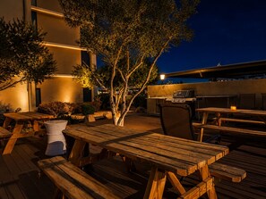 An outdoor patio with wooden tables and chairs, a barbecue grill, and a tree.