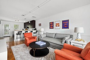 A modern living room with a grey sofa, orange armchairs, a black coffee table, and a kitchen in the background. - Royal Tiger - Walk to Death Valley! (Clemson)