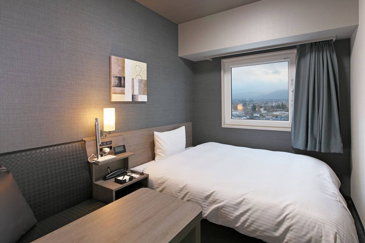 Habitación de hotel con cama, escritorio, mesita de noche, lámpara, reloj y ventana con vista a la ciudad.