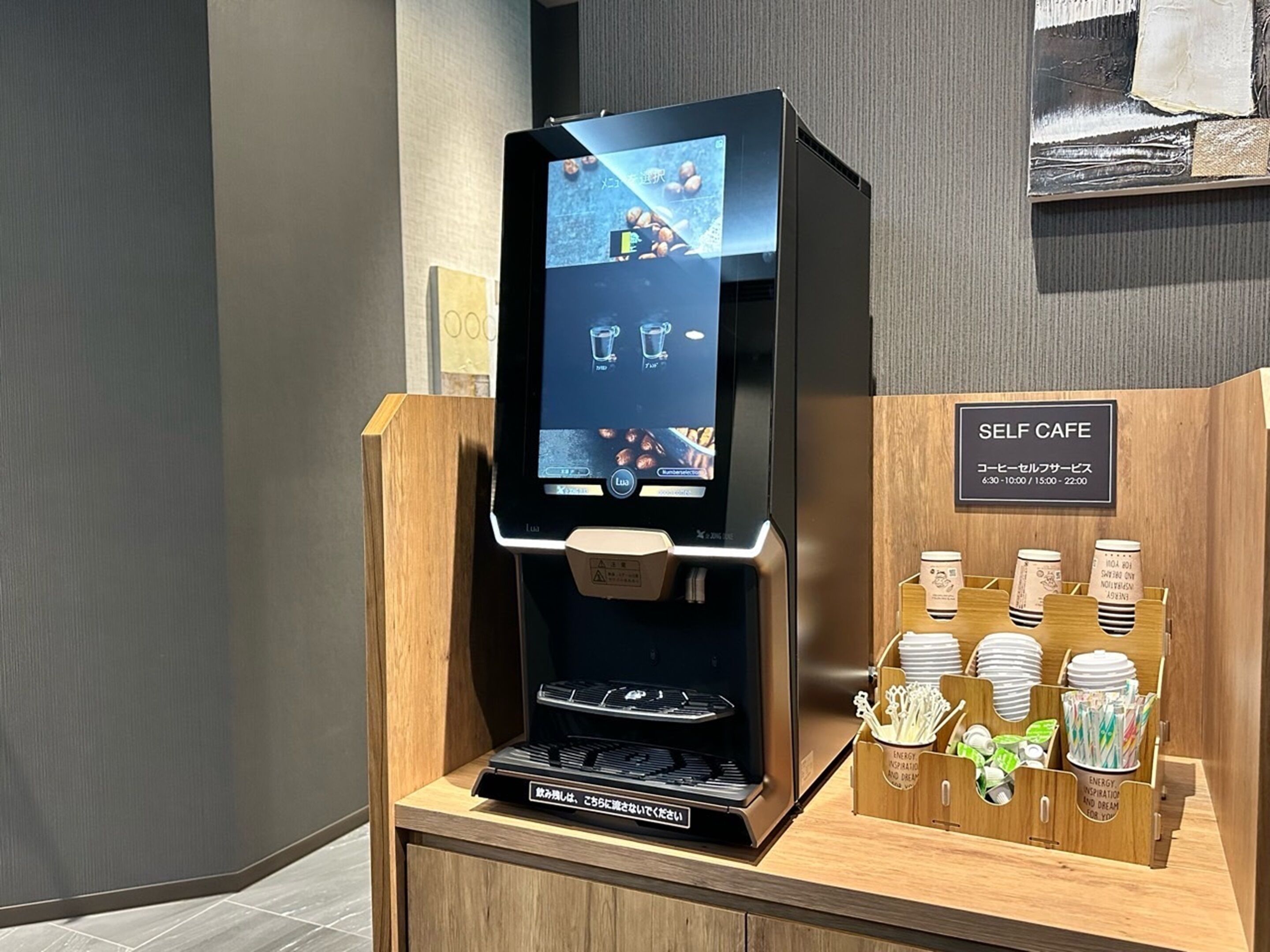 A self-service coffee machine with a digital interface, a coffee dispenser, and a display of disposable cups and utensils.