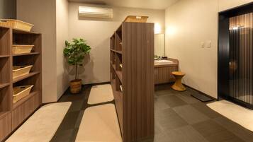 A hotel room with a wooden floor, a large mirror, a sink, a plant, and a shelf with baskets.