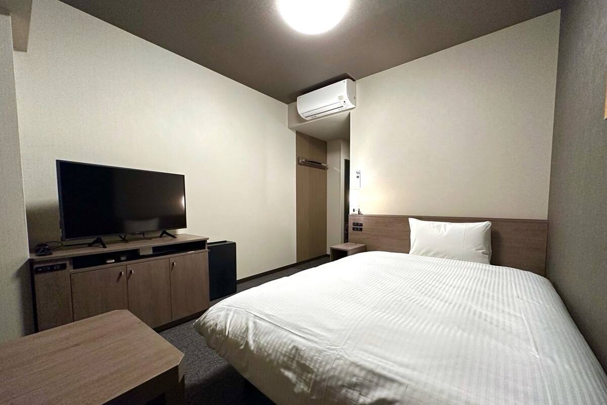A hotel room with a bed, a TV on a wooden cabinet, a desk, and an air conditioning unit on the wall.