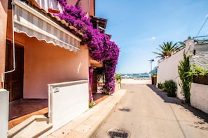Eine enge Straße mit einem Gebäude, das einen Balkon mit violetten Blumen, einen klaren Himmel und einen weiten Blick auf das Meer zeigt.