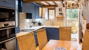 A kitchen with blue cabinets, a wooden table, wicker chairs, and a hanging light fixture. - Granary Cottage - Luxury Barn Conversion (Chipping Campden)