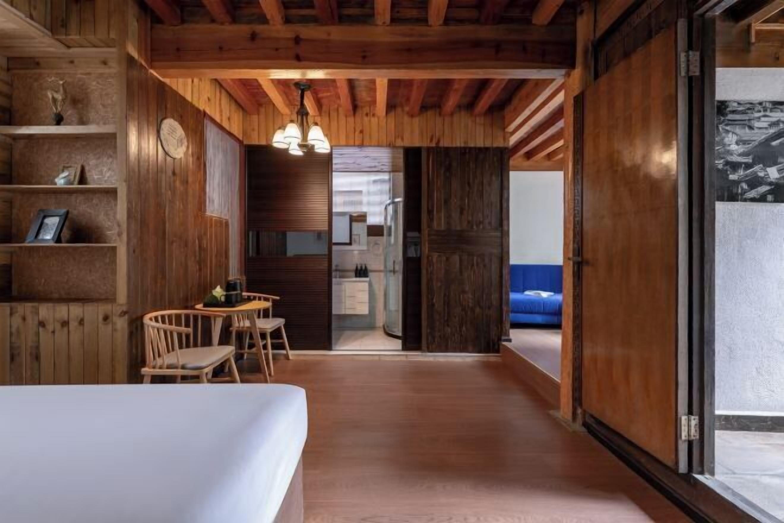 Interior em madeira com uma cama, uma escrivaninha e uma cadeira. Há uma porta que dá para outro quarto.