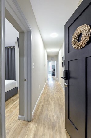 A hallway with a bed, a dark door with a woven decoration, and a kitchen in the background. - Parkside Gem: Lux & Spacious-Historic Druid Hill (Baltimore)