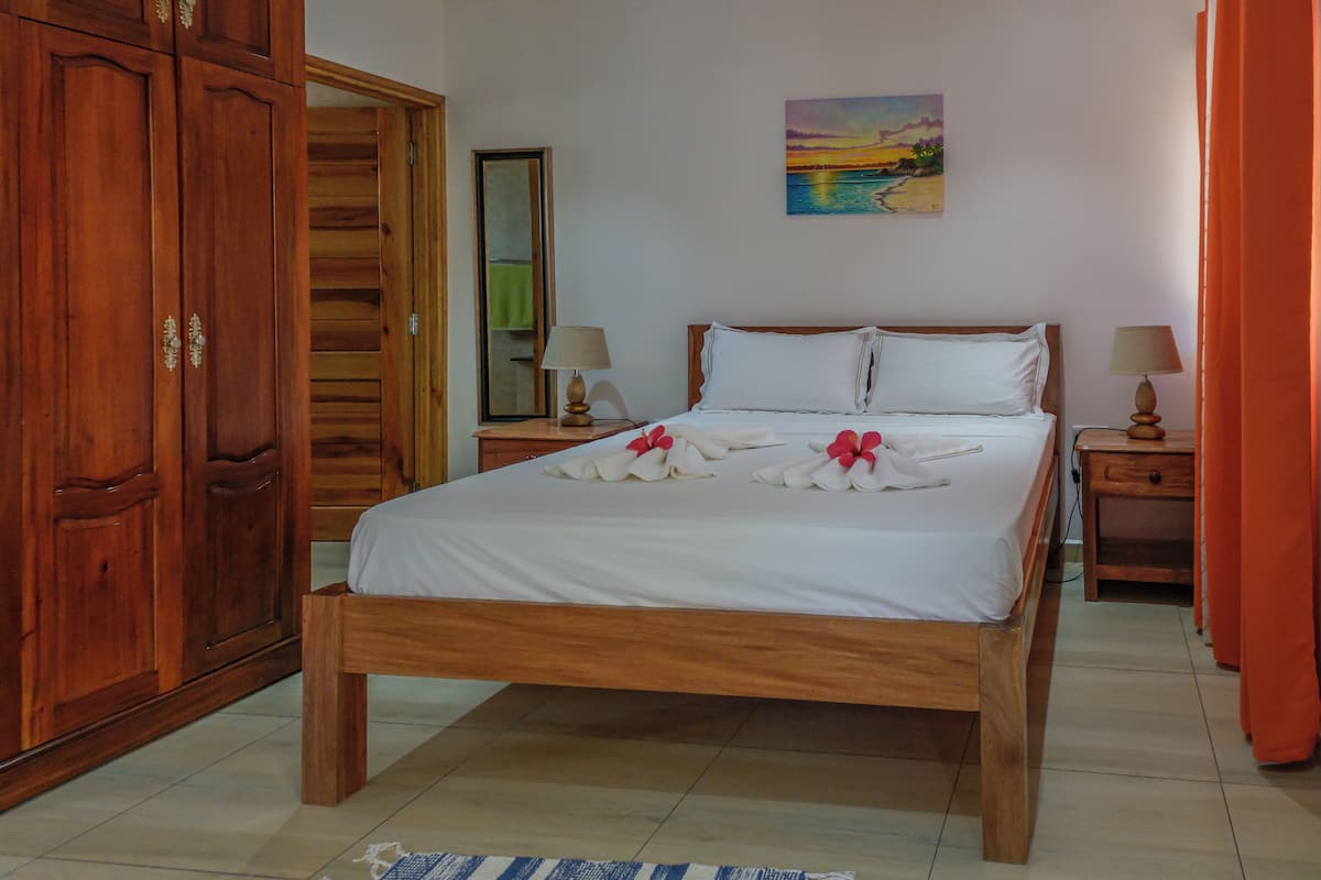 Una cama bien hecha con sábanas blancas y arreglos florales rojos, muebles de madera y un cuadro en la pared.