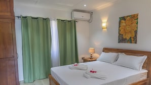 Habitación de hotel con una cama de madera, cortinas verdes, aire acondicionado de pared, una lámpara de noche y un cuadro con motivos florales.