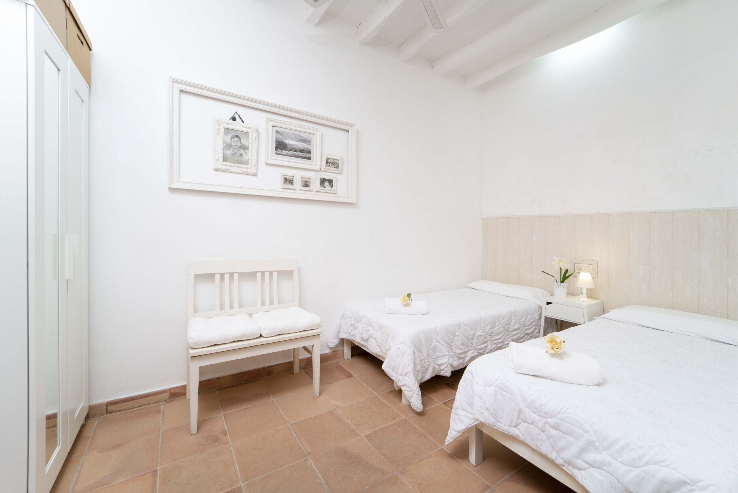 A clean, white room with two beds, a bench, and a wall-mounted picture frame.