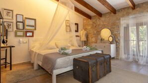 Une chambre à coucher avec un lit, une table de chevet, une valise, un mur en pierre et une fenêtre avec des rideaux légers.