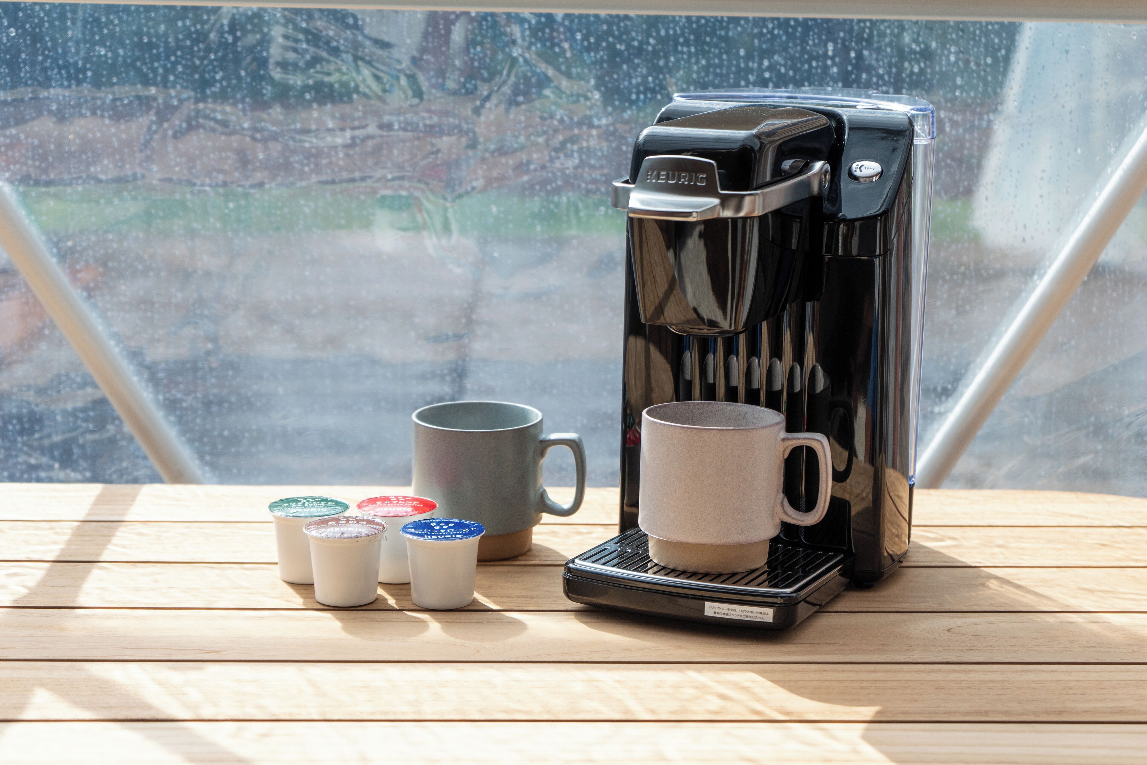 Una máquina de café Keurig sobre una mesa de madera con una taza gris y tres tazas pequeñas con tapa.