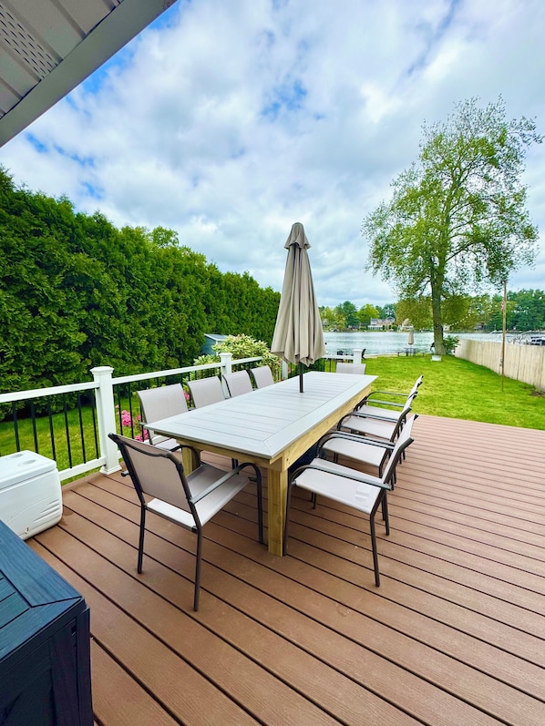 Eine Terrasse im Freien mit einem Holztisch und Stühlen, umgeben von einem weißen Geländer, mit Blick auf einen See und Grünflächen.