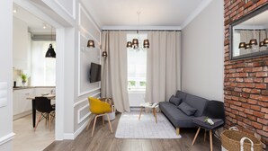A modern living room with a grey sofa, a yellow chair, and a white rug. There is a TV mounted on the wall and a brick wall on one side.