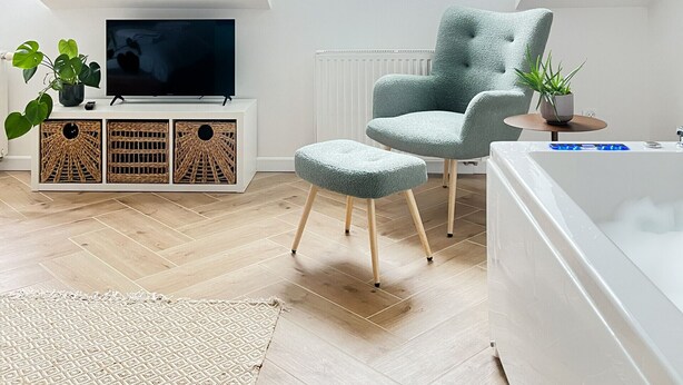 Un salon moderne avec un plafond en pente, une grande fenêtre, un téléviseur, un fauteuil confortable, un petit pouf, un meuble blanc avec des paniers en osier et une baignoire blanche.