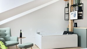 Une chambre moderne avec une baignoire blanche, un fauteuil vert et une petite table ronde sur laquelle se trouve une bouteille.