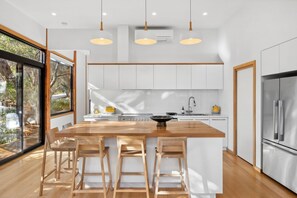 A modern kitchen with a central island, wooden bar stools, pendant lighting, and a large window offering a view of the outdoors. - Carji (Anglesea)