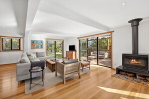 A modern living room with a fireplace, wooden flooring, and a large sliding glass door leading to an outdoor patio. - Carji (Anglesea)
