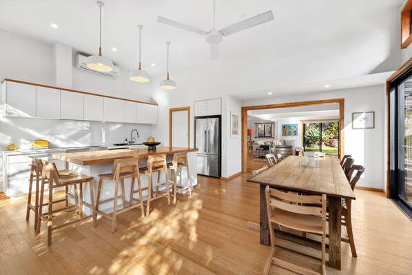 A modern kitchen with white cabinets, a wooden island with bar stools, and a dining area with a large wooden table and chairs. - Carji (Anglesea)