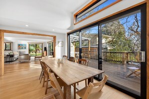 A spacious dining area with a large wooden table and chairs, a view of the outdoors through large sliding glass doors, and a cozy living area with a sofa and artwork. - Carji (Anglesea)