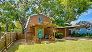 Une cabane en bois avec un porche, un espace abrité et une maison bleue en arrière-plan.