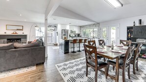 A spacious kitchen with a central island, a dining area with wooden chairs and a patterned rug, and a living area with a sofa and a fireplace.