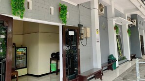 Un pasillo con ventilador de techo, unidades de aire acondicionado y plantas verdes. Hay bancos a lo largo del pasillo y un basurero.