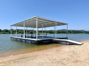 A covered dock with a bench and a table, extending into a lake with a sandy beach and hills in the background.