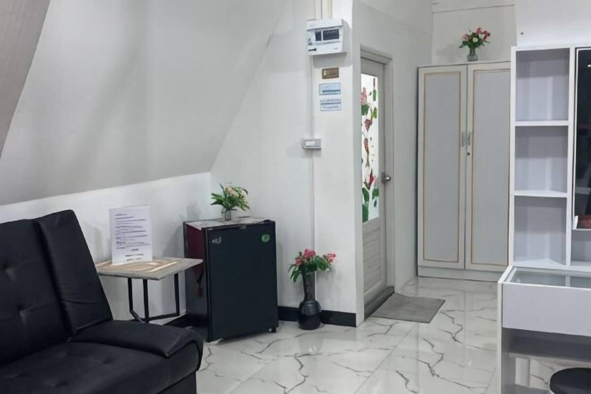 A reception area with a black sofa, a small table with a plant, a refrigerator, a door with a sign, and a white cabinet with shelves.