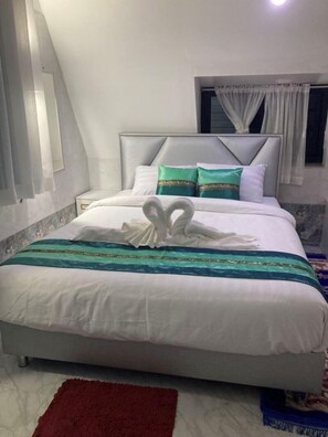 Cama bem arrumada com arranjo de toalhas em formato de coração, cabeceira e janela com cortinas.
