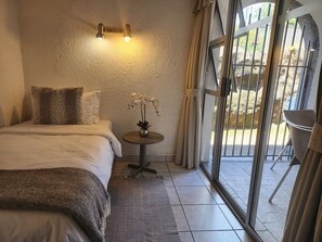 A bedroom with a bed, a small table with a vase, a window with curtains, and a view of the outside.