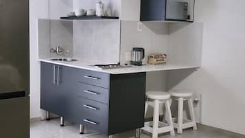 A compact kitchen with a built-in sink, a coffee maker, and a microwave. There are two stools and a shelf with kitchenware.