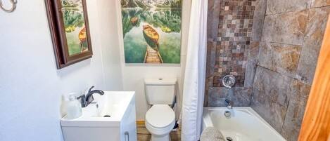 Ein Badezimmer mit Holzboden, Waschbecken, Toilette und Badewanne. An der Wand hängen zwei gerahmte Bilder.