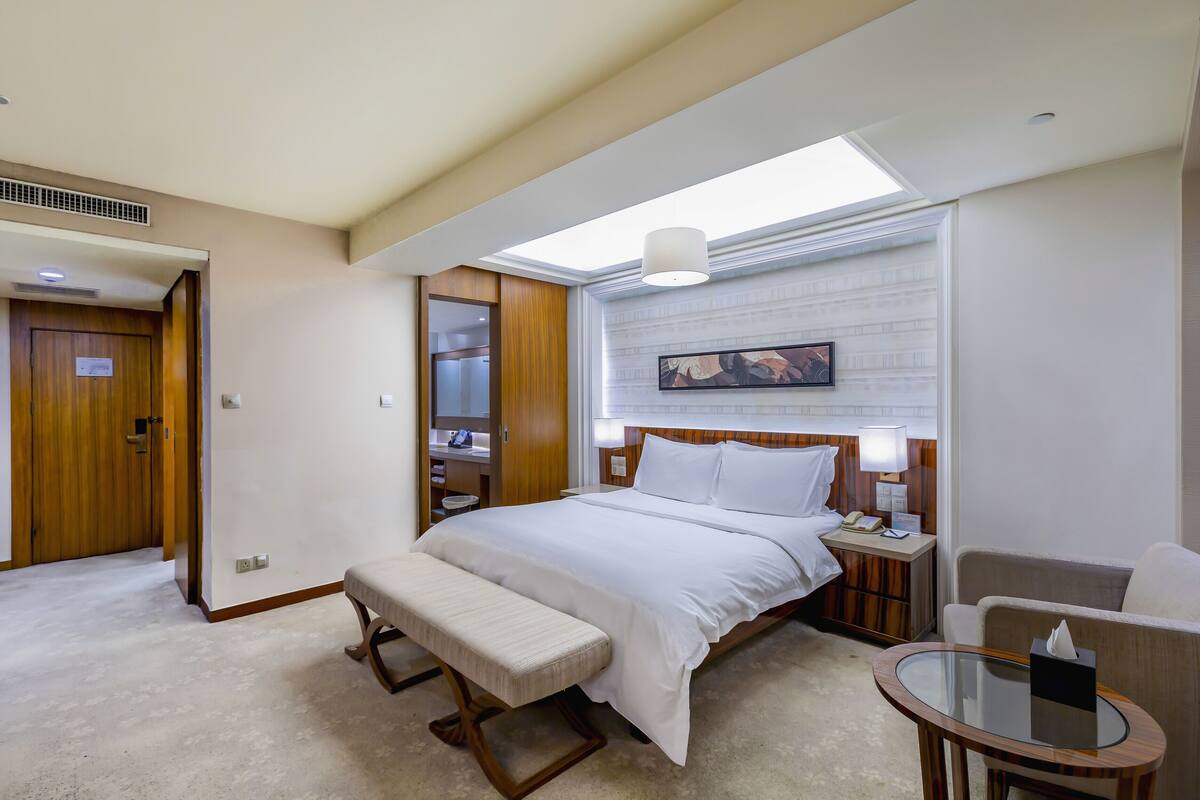 Een hotelkamer met een groot bed, een bankje, een nachtkastje met een lamp, een stoel en een tafeltje met een zakdoos.
